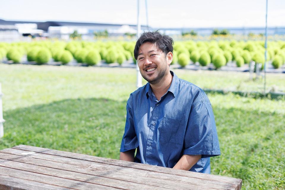 写真：緑の自然を背景に、青い半袖シャツを着た男性がインタビューを笑顔で受けている様子