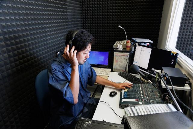写真：ラジオ局の機材を前に、ヘッドフォンをして収録音声を確認している男性